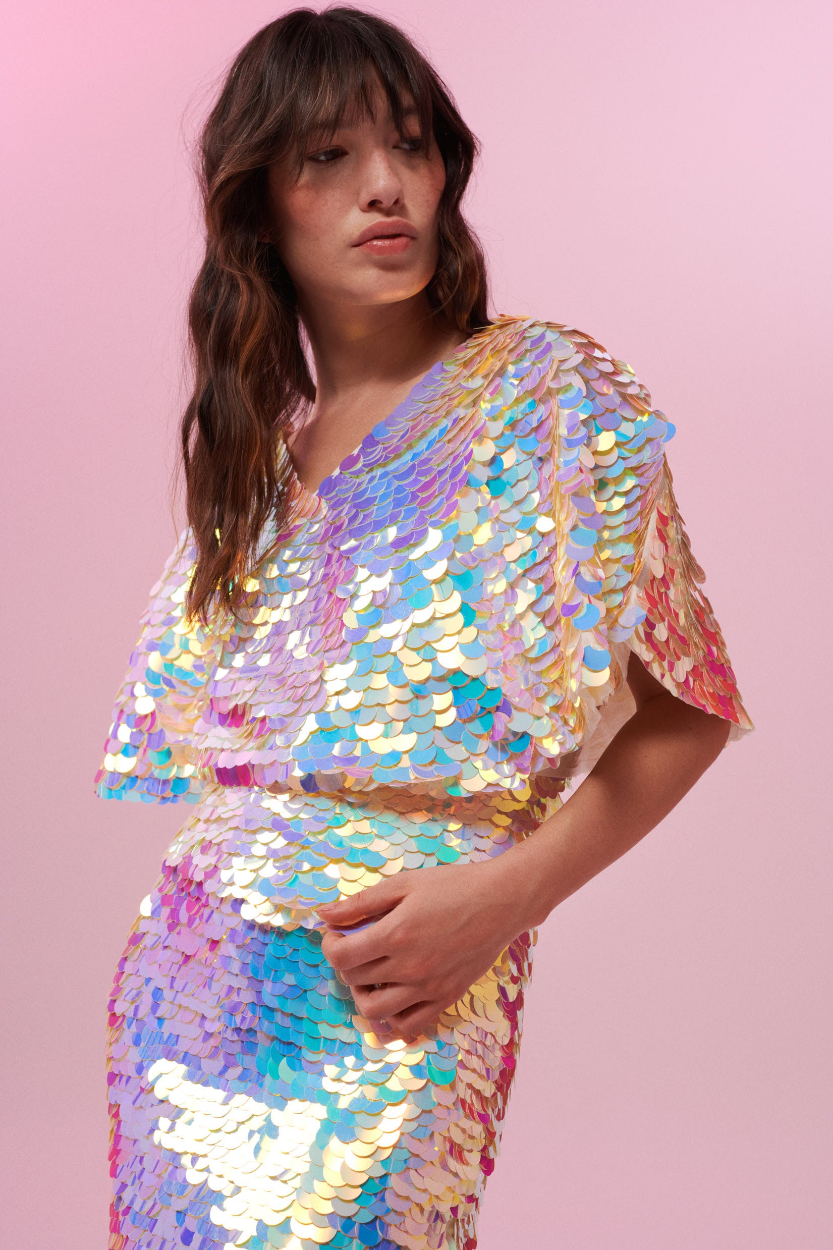 A woman facing to the side at an angle wearing an opal sequin top with large cape sleeves covered in large round holographic purple, pink, blue and white coloured Rosa Bloom sequins.