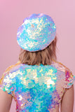 Rear view of a woman wearing a sequin beret made from small round holographic sequins with hues of lilac purple, light blue and soft pinks. The festival sequin hat matches the sequin top that the model is wearing. The Opal sequins by Rosa Bloom glisten, creating a mix of shimmering colours.  