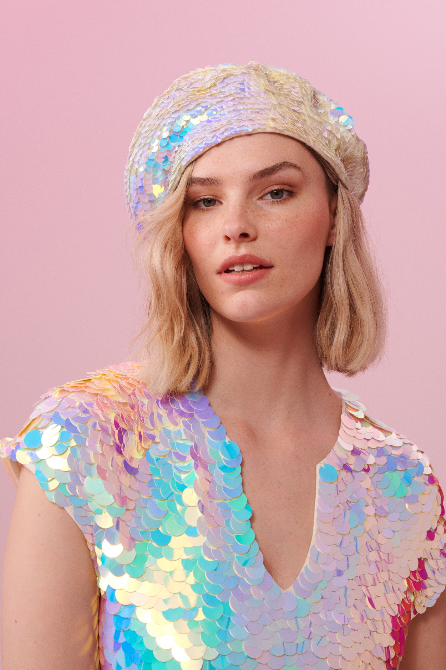 A woman with short blonde hair, wearing a Grace sequin beret made from small round holographic sequins with hues of lilac purple, light blue and soft pinks. The festival sequin hat matches the sequin top that the model is wearing. The Opal sequins by Rosa Bloom glisten, creating a mix of shimmering colours.  