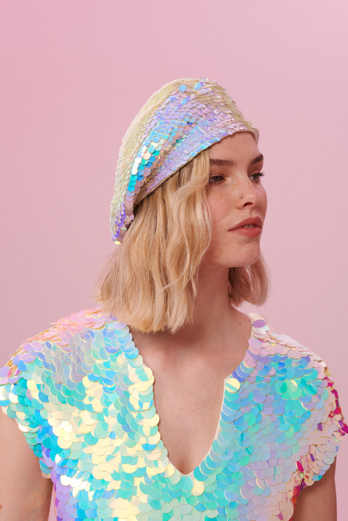 A side on view of a woman with short blonde hair, wearing a Grace sequin beret made from small round holographic sequins with hues of lilac purple, light blue and soft pinks. The festival sequin hat matches the sequin top that the model is wearing. The Opal sequins by Rosa Bloom glisten, creating a mix of shimmering colours.  