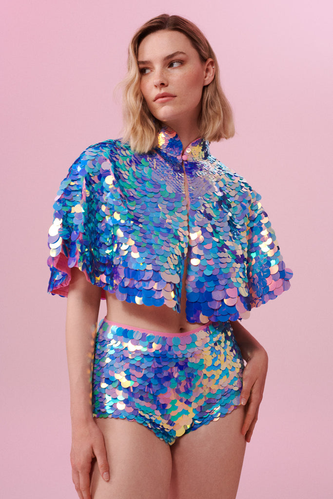  A woman with short blonde hair, standing face on, but slightly to the side, wearing high waisted  sequin hot pant shorts and a cape completely covered in large round holographic Rosa Bloom sequins. The sequins glisten in the light, creating a mix of shimmering colours of pink, blue and lilac. 