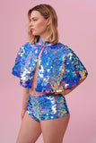 A woman with short blonde hair, standing face on, but slightly to the side with her hand on her hip,  wearing, high cut sequin hot pant shorts and a cape completely covered in large round holographic Rosa Bloom sequins. The sequins glisten in the light, creating a mix of shimmering colours of pink, blue and lilac. 