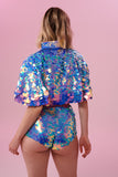  The back of a woman with short blonde hair, wearing high waisted, high cut sequin hot pant shorts and a cape completely covered in large round holographic Rosa Bloom sequins. The sequins glisten in the light, creating a mix of shimmering colours of pink, blue and lilac. 