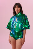 A woman long brown hair with a fringe, wearing stretchy high waisted  Gigi festival sequin hot pants shorts and a matching sequin cape completely covered in large round holographic Rosa Bloom sequins. The Emerald  sequins sparkle in the light, creating a mix of shimmering colours of emerald green and ultramarine blue.