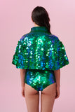 A woman long brown hair wearing stretchy high cut Gigi festival sequin hot pants shorts and a matching sequin cape completely covered in large round holographic Rosa Bloom sequins. The Emerald  sequins sparkle in the light, creating a mix of shimmering colours of emerald green and ultramarine blue.