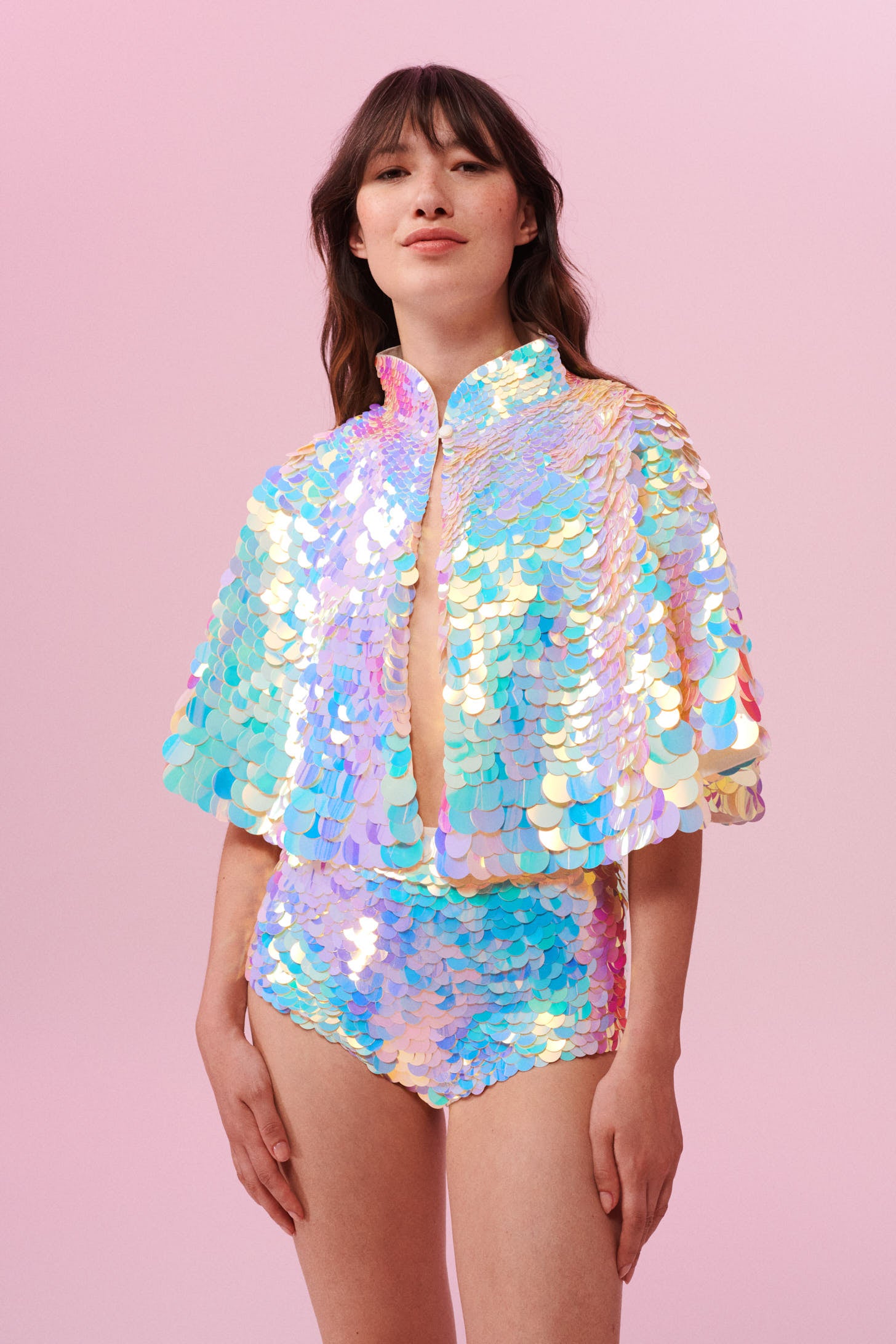 A woman long brown hair with a fringe, swept back wearing an opal sequin high knecked cape and short sequin hot pant shorts covered in large round holographic purple, pink, blue and white coloured Rosa Bloom sequins. The sequins glisten, creating a mix of shimmering colours of pink, blue, lilac and white.