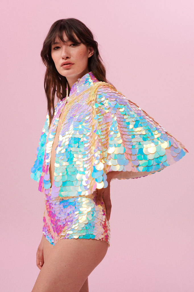 A woman long brown hair with a fringe, swept back to the side, wearing an opal sequin high knecked cape with her hand on her hip, with short sequin hot pant shorts covered in large round holographic purple, pink, blue and white coloured Rosa Bloom sequins. The sequins glisten, creating a mix of shimmering colours of pink, blue, lilac and white. 