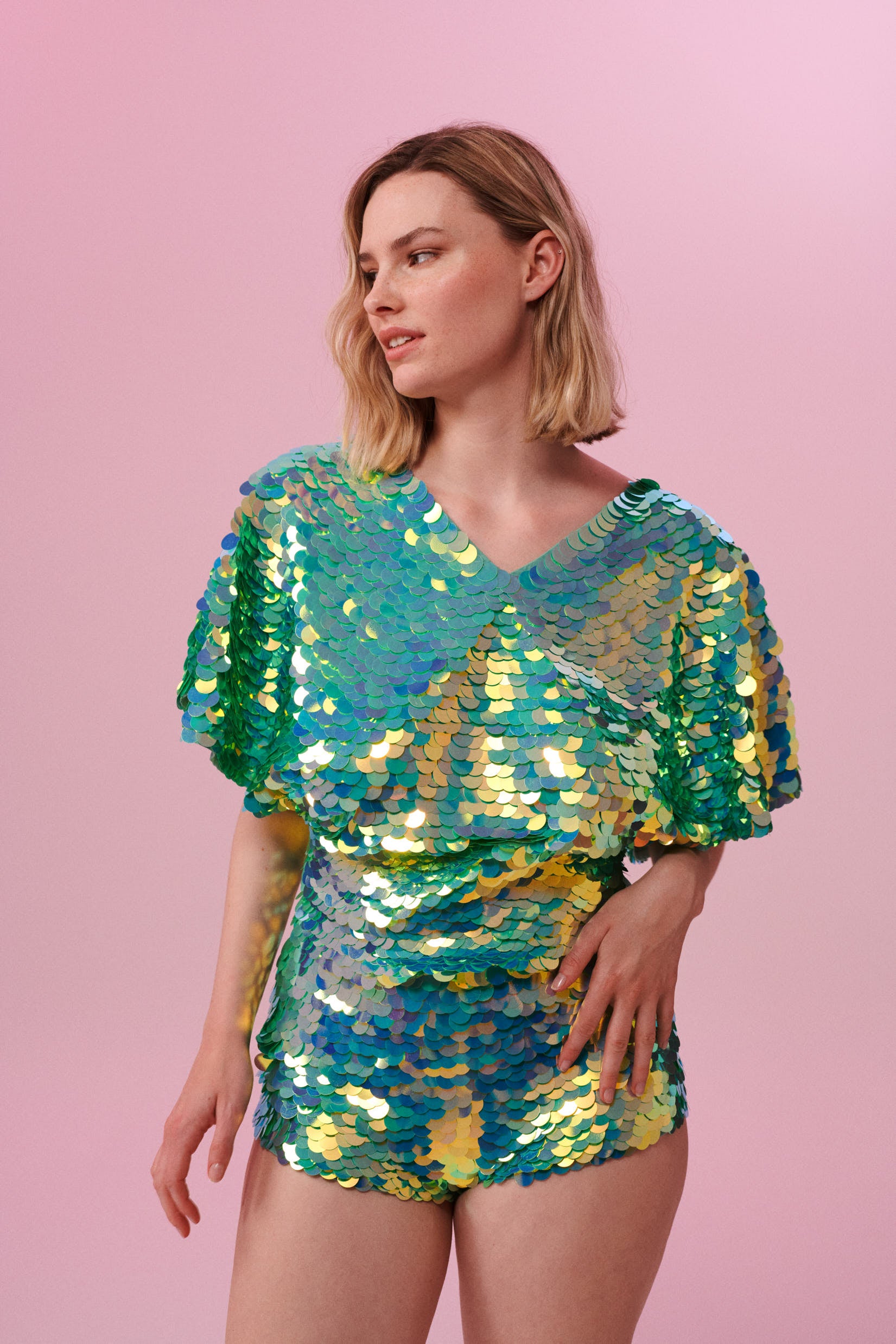 A woman with short blonde hair, wearing high waisted  festival sequin hot pants shorts and a sequin cape completely covered in large round holographic Rosa Bloom sequins. The Chameleon sequins glisten in the light, creating a mix of shimmering colours of sage green, lilac and mint. 