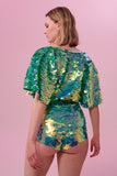 A woman with short blonde hair, standing with her back to you, looking slightly to the side, wearing high waisted  festival sequin hot pants shorts and a cape completely covered in large round holographic Rosa Bloom sequins. The Chameleon sequins glisten in the light, creating a mix of shimmering colours of sage green, and mint. 