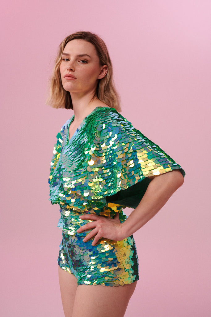 Blonde haired model posing side on wearing Rosa Bloom pale green sequinned cape top