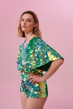 A woman with short blonde hair, wearing high waisted  festival sequin hot pants shorts and a sequin cape completely covered in large round holographic Rosa Bloom sequins. The Chameleon sequins glisten in the light, creating a mix of shimmering colours of sage green, lilac and mint. 