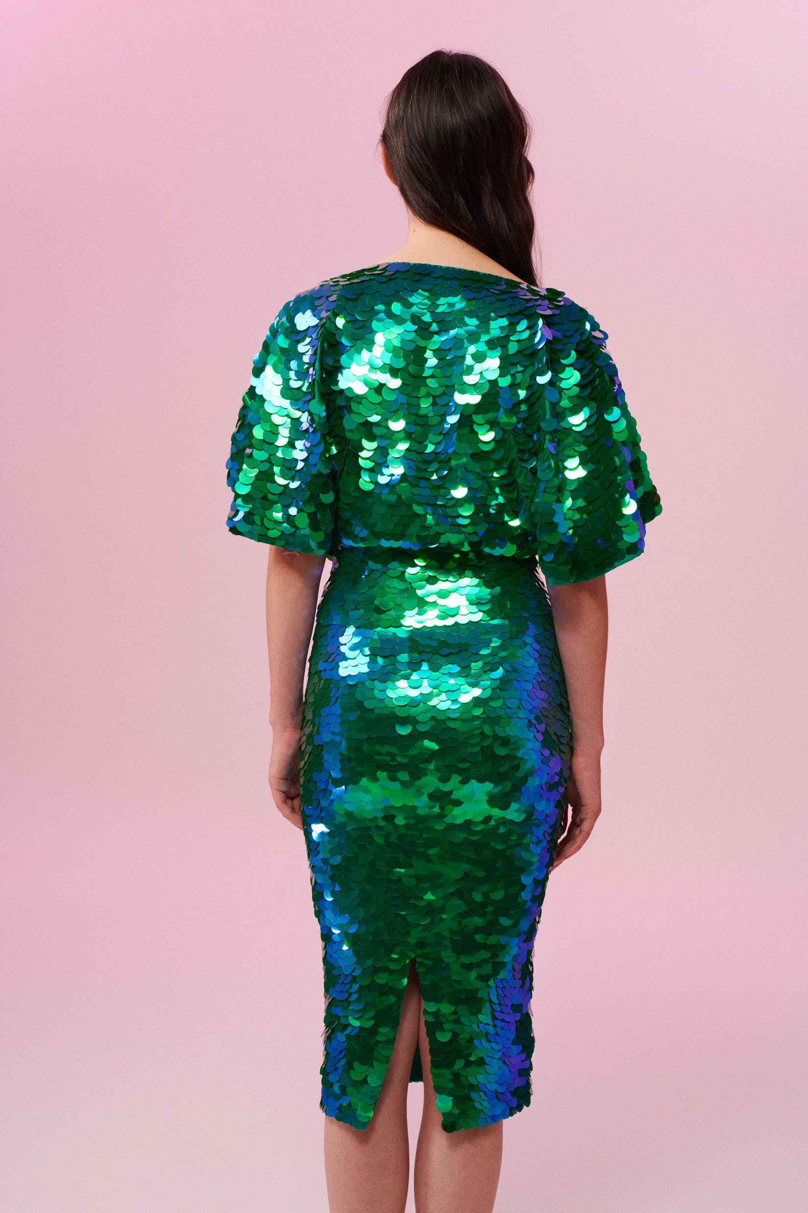 Rear view of a dark haired woman wearing a Rosa Bloom emerald green sequin mid length sequin tube skirt