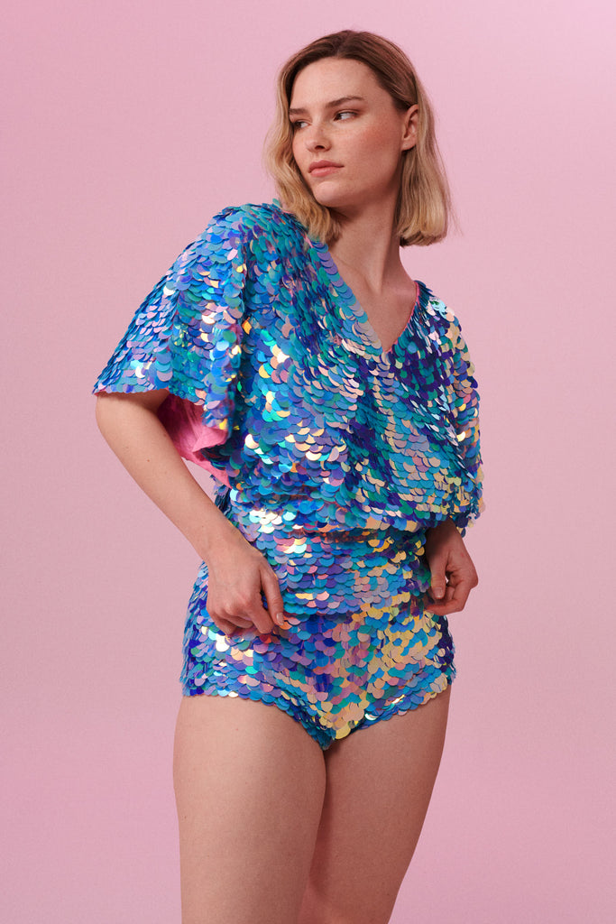 Blonde haired model posing side on wearing Rosa Bloom blue and pink sequinned cape top