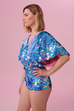Blonde haired model posing side on wearing Rosa Bloom blue and pink sequinned cape top