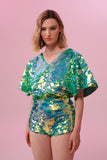 A woman with short blonde hair, wearing high waisted  festival sequin hot pants shorts and a sequin cape completely covered in large round holographic Rosa Bloom sequins. The Chameleon sequins glisten in the light, creating a mix of shimmering colours of sage green, lilac and mint. 