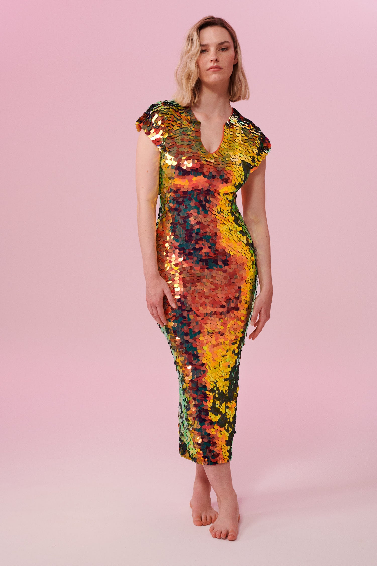 A woman with short blonde hair standing facing the viewer wearing an iridescent golden sequin long dress. A figure hugging  dress down to the ankles with small capped sleeves completely covered in large round holographic Rosa Bloom sequins. The sequins glisten, creating a mix of shimmering colours of burnt orange, yellow, gold and warm red making this sequin dress glow when worn.