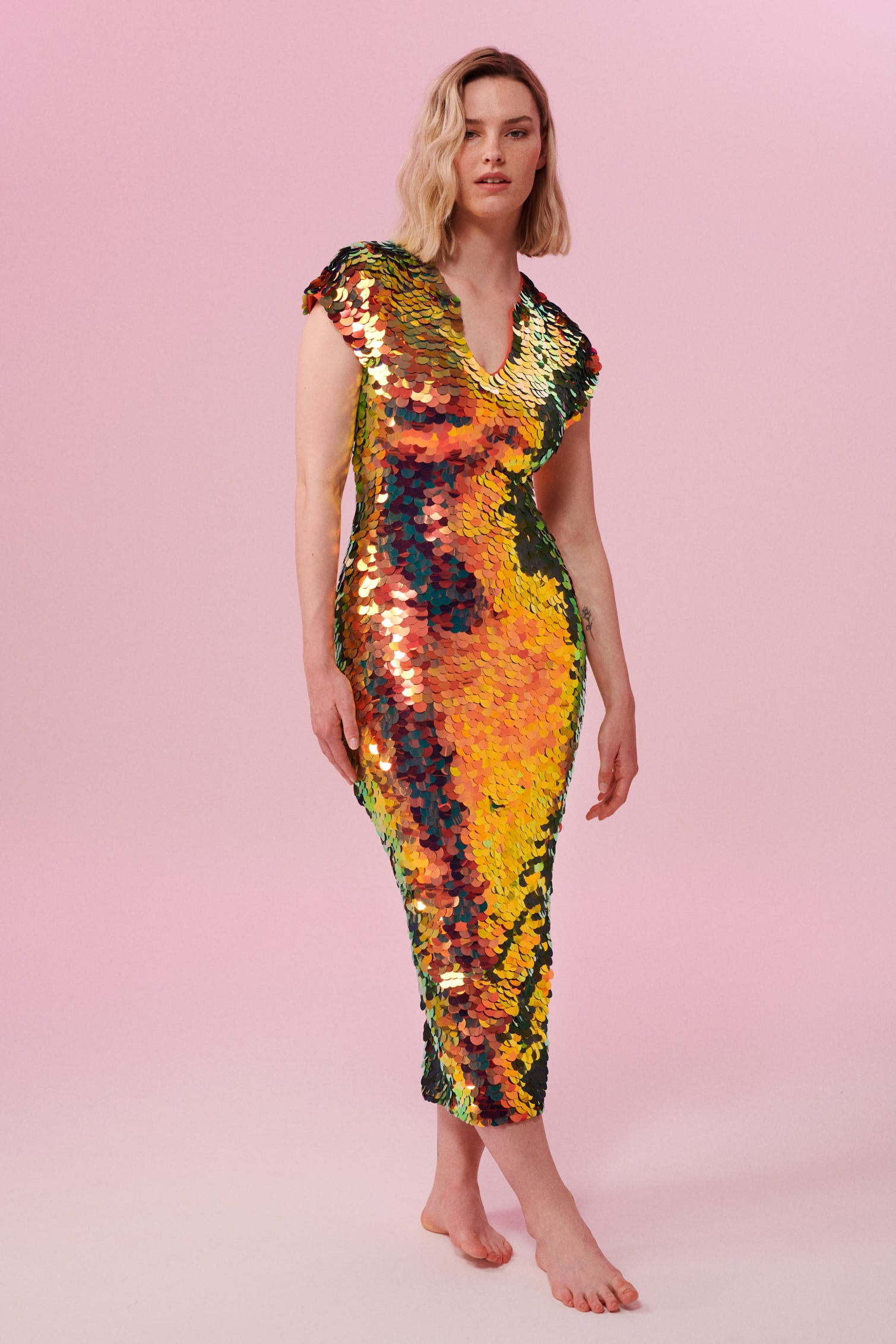 A woman with short blonde hair standing facing the viewer wearing an iridescent golden sequin long dress. A figure hugging  dress down to the ankles with small capped sleeves completely covered in large round holographic Rosa Bloom sequins. The sequins glisten, creating a mix of shimmering colours of burnt orange, yellow, gold and warm red making this sequin dress glow when worn.