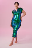 A woman with tied back brown hair standing facing the viewer wearing an iridescent green and blue sequin long dress. A figure hugging  long dress with small capped sleeves completely covered in large round holographic Rosa Bloom sequins. The sequins glisten, creating a mix of shimmering colours of emerald green and ultramarine blue, this sequin dress looks like it is glowing when worn. 