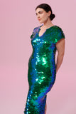 A woman with brown hair tied back in a bun, standing slightly to the side wearing an iridescent green and blue sequin long dress. A figure hugging long sequin dress with small capped sleeves,completely covered in large round holographic Rosa Bloom sequins. The sequins glisten, creating a mix of shimmering colours of emerald green and ultramarine blue, this sequin dress looks like it is glowing  when worn. 