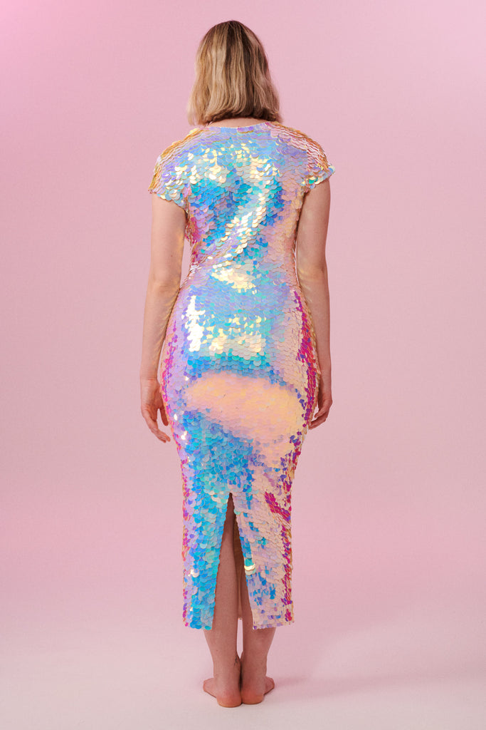 A woman with short blonde hair with her back to us wearing an opal sequin long figure hugging dress with small capped sleeves covered in large round holographic purple, pink, blue and white coloured Rosa Bloom sequins. The sequins glisten, creating a mix of shimmering colours of pink, blue, lilac and white. 