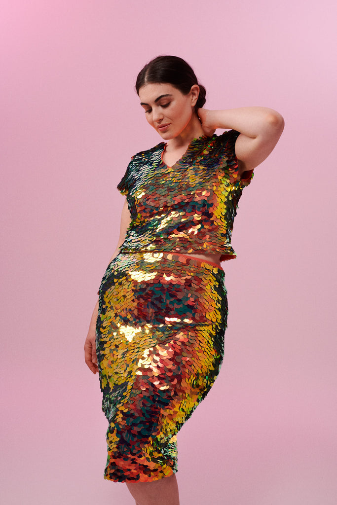 A woman with brown hair, wearing a sequin stretchy top made with large round holographic Rosa Bloom sequins. The ember sequins in this Gwen design glistens in the light, creating a mix of shimmering colours of burnt orange, gold, red and orange. The model is also wearing a matching sequin skirt. 