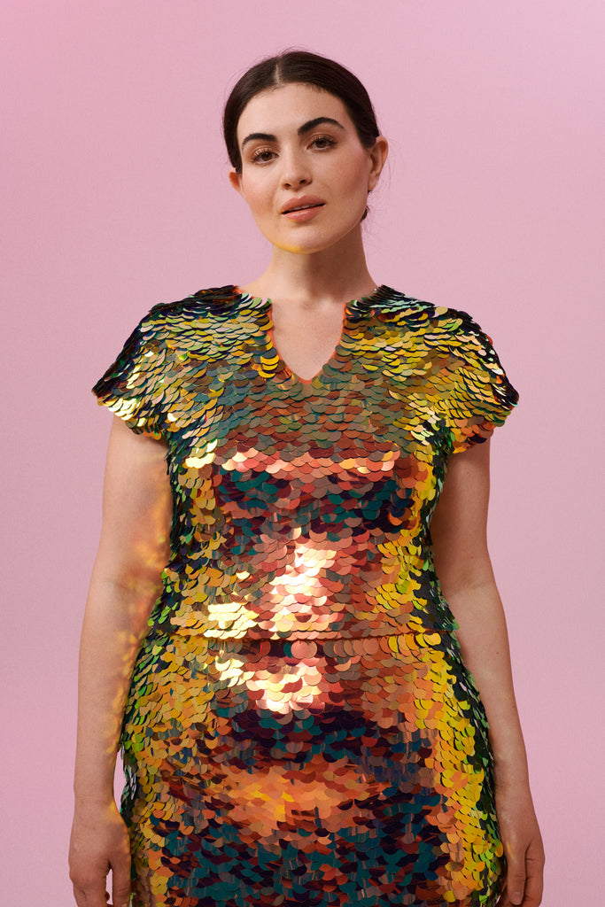 A woman with brown hair, wearing a sequin stretchy top made with large round holographic Rosa Bloom sequins. The ember sequins in this Gwen design glistens in the light, creating a mix of shimmering colours of burnt orange, gold, red and orange. The model is also wearing a matching sequin skirt. 