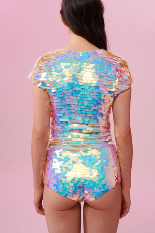 A rear view of a woman with brown hair, wearing a sequin stretchy top with capped sleeves made with large round holographic Rosa Bloom sequins. The Gwen sequins glisten, creating a mix of shimmering colours of soft gold, aqua, lilac, and peach. The model is also wearing matching high waisted, stretchy festival sequin shorts in the same matching sequin colour way.