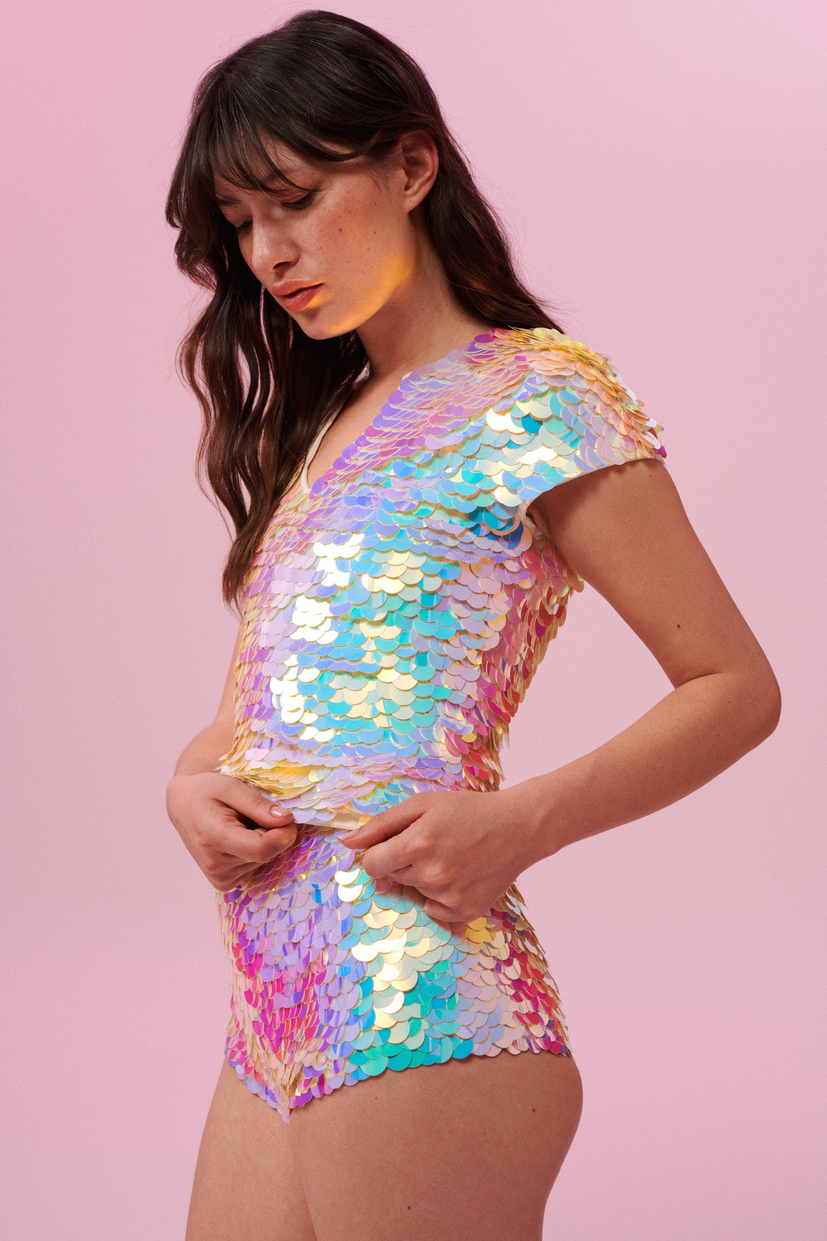 A side on view of a woman with brown hair, wearing a sequin stretchy top with capped sleeves made with large round holographic Rosa Bloom sequins. The Gwen sequins glisten, creating a mix of shimmering colours of soft gold, aqua, lilac, and peach. The model is also wearing matching high waisted, stretchy festival sequin shorts in the same matching sequin colour way.