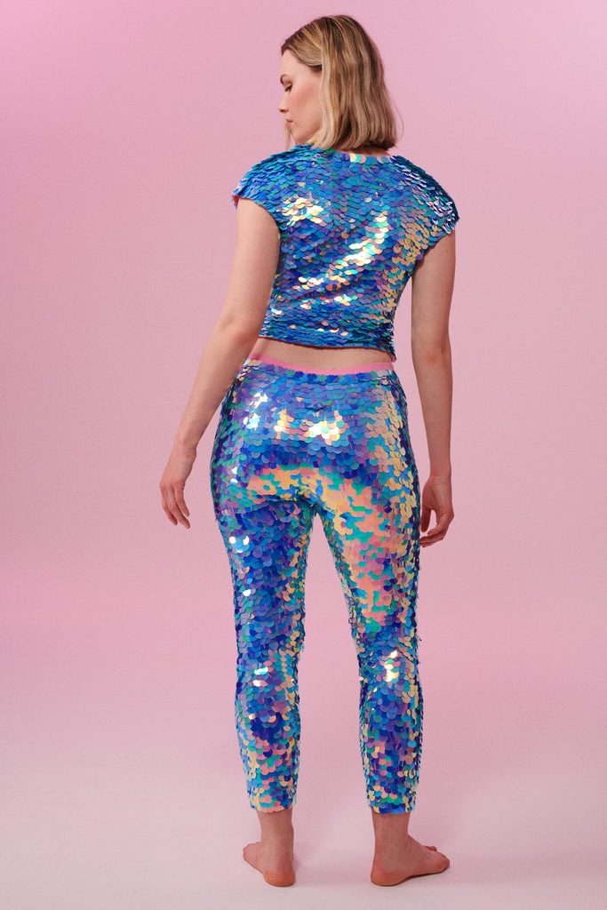A rear view of a woman wearing iridescent pink, purple and blue, stretchy sequin Indus leggings covered in large round holographic Rosa Bloom sequins. The model is also wearing a matching stretchy sequin top, in matching colours. The sequins glisten, creating a mix of shimmering colours  to create this Amethyst sequin design that looks like it is glowing. 