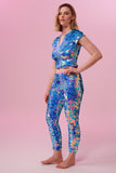 A woman wearing iridescent pink, purple and blue, stretchy sequin Indus leggings covered in large round holographic Rosa Bloom sequins. The model is also wearing a matching stretchy sequin top, in matching colours. The sequins glisten, creating a mix of shimmering colours  to create this Amethyst sequin design that looks like it is glowing. 