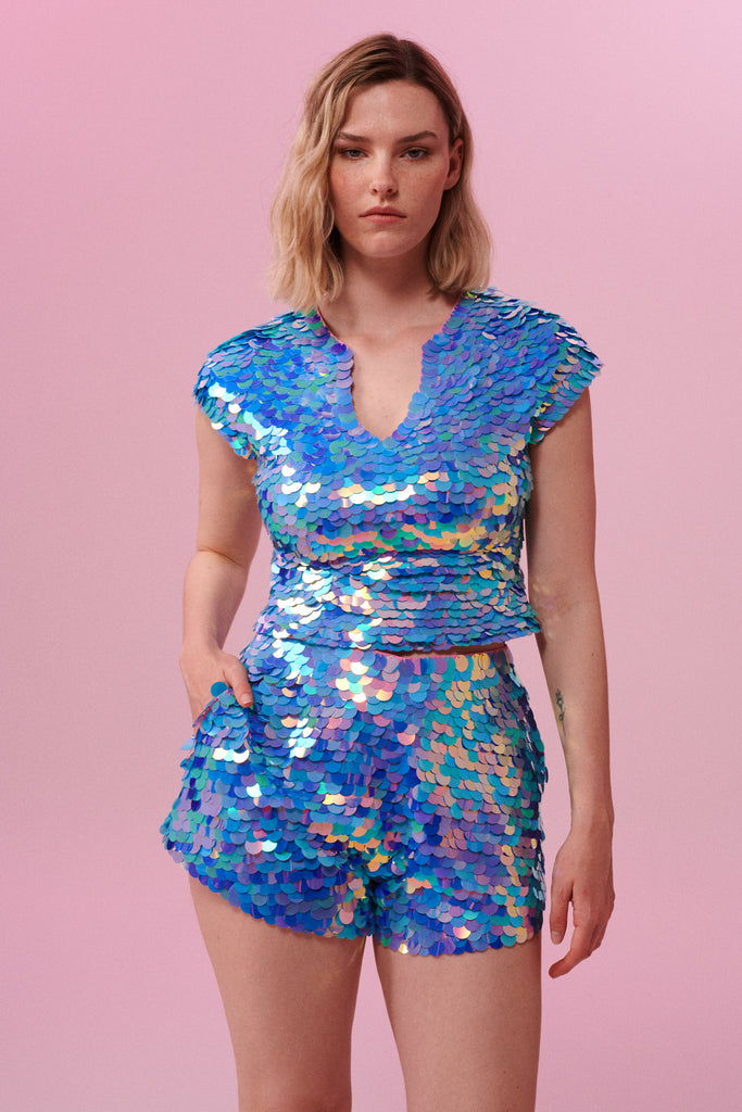 A woman wearing iridescent purple, pink, blue and white Juno high waisted sequin shorts covered in large round holographic Rosa Bloom sequins. The sequins glisten, creating a mix of shimmering colours make this sequin shimmer. The model is also wearing a matching stretchy sequin top, in matching colours. This Amethyst sequin outfit looks like it is glowing in the light. 