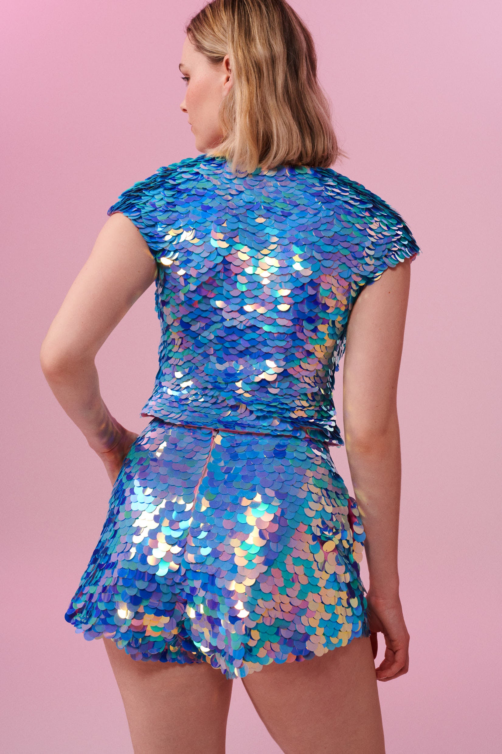 A rear view of a woman with short blonde hair, wearing high waisted festival sequin shorts and a matching sequin top made with large round holographic Rosa Bloom sequins. The Amethyst  sequins glisten in the light, creating a mix of shimmering colours of lilac purple, light blue and soft pinks. 