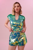 A front view of a woman with short blonde hair, wearing high waisted festival sequin shorts and a matching sequin stretchy top made with large round holographic Rosa Bloom sequins. The chameleon sequins in this Gwen design glistens in the light, creating a mix of shimmering colours of sage and mint greens. 