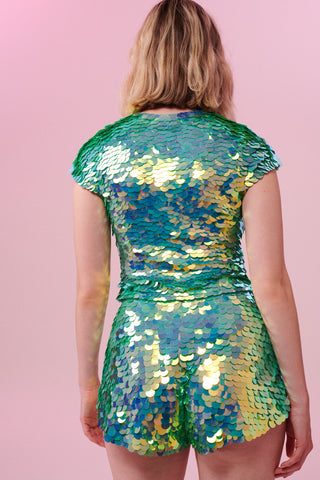 A rear view of a woman with short blonde hair, wearing high waisted festival sequin shorts and a matching sequin stretchy top made with large round holographic Rosa Bloom sequins. The chameleon sequins in this Gwen design glistens in the light, creating a mix of shimmering colours of sage and mint greens. 