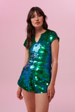 A woman wearing iridescent green and blue Juno high waisted, tailored sequin shorts covered in large round holographic Rosa Bloom sequins. The sequins glisten, creating a mix of shimmering colours of greens and blues. The model is also wearing a matching stretchy sequin cropped vest, in matching colours.