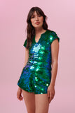 A woman with brown hair, wearing a sequin stretchy top with capped sleeves made with large round holographic Rosa Bloom sequins. The Gwen sequins glisten, creating a mix of shimmering colours of emerald green and ultramarine blue. The model is also wearing matching festival sequin high waisted shorts. 