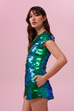A side view of a woman with brown hair, wearing a sequin stretchy top with capped sleeves made with large round holographic Rosa Bloom sequins. The Gwen sequins glisten, creating a mix of shimmering colours of emerald green and ultramarine blue. The model is also wearing matching festival sequin shorts. 