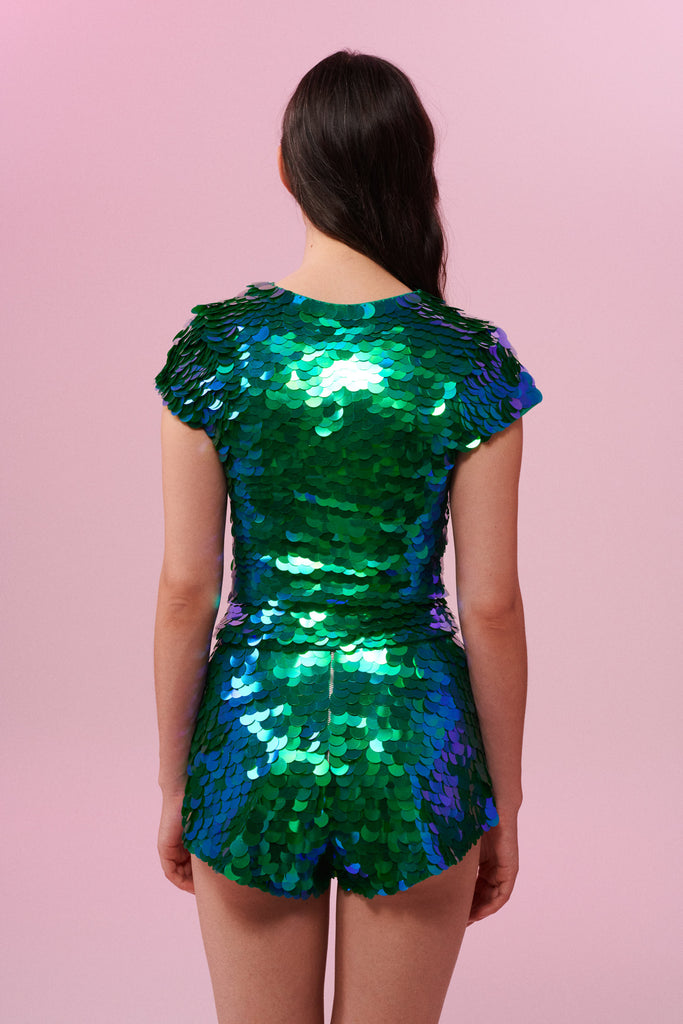 A woman standing with her back to the viewer wearing iridescent green and blue Juno high waisted, tailored sequin shorts covered in large round holographic Rosa Bloom sequins. The sequins glisten, creating a mix of shimmering colours of greens and blues. The model is also wearing a matching stretchy sequin cropped vest, in matching colours. 