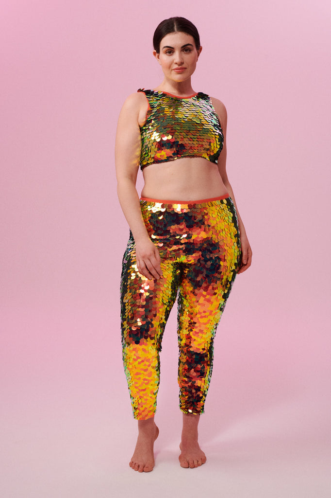 A woman wearing iridescent yellow, red and orange stretchy Indus leggings covered in large round holographic Rosa Bloom sequins. The sequins glisten, creating a mix of shimmering colours make this sequin glow. The model is also wearing a matching stretchy sequin vest top, in matching colours. This Ember sequin outfit looks like it is glowing. 