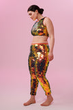 A woman wearing iridescent yellow, red and orange stretchy Indus leggings covered in large round holographic Rosa Bloom sequins. The sequins glisten, creating a mix of shimmering colours make this sequin glow. The model is also wearing a matching stretchy sequin vest top, in matching colours. This Ember sequin outfit looks like it is glowing. 