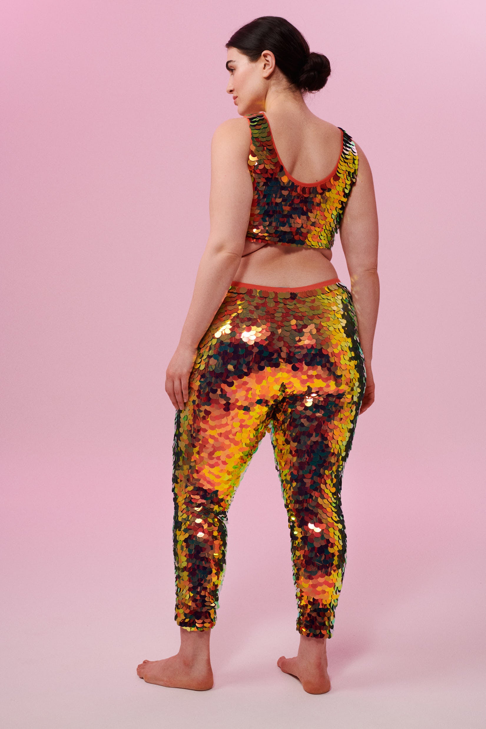 A rear view of a woman wearing iridescent yellow, red and orange stretchy Indus leggings covered in large round holographic Rosa Bloom sequins. The sequins glisten, creating a mix of shimmering colours make this sequin glow. The model is also wearing a matching stretchy sequin vest top, in matching colours. This Ember sequin outfit looks like it is glowing. 