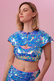 A woman with short blonde hair, wearing a festival sequin cape made with small and large round holographic Rosa Bloom sequins. The Amethyst sequins, sitting neatly over her shoulders in this Inti design glistens in the light, creating a mix of shimmering colours of purple, pink, blue and white. The model is also wearing a matching sequin vest top and sparkly leggings.