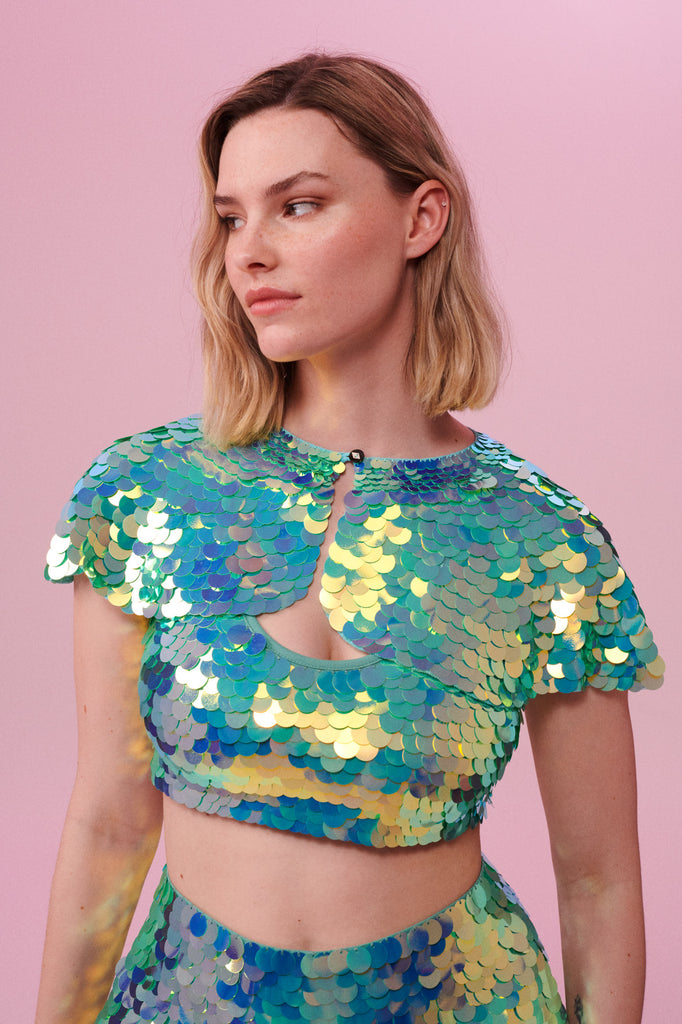 A woman with short blonde hair, wearing a festival sequin cape made with small and large round holographic Rosa Bloom sequins. The Chameleon sequins, sitting neatly over her shoulders in this Inti design glistens in the light, creating a mix of shimmering colours of pale green sequinned mint and warm peachy gold. The model is also wearing a matching sequin vest top and sparkly leggings.