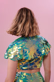 A woman with short blonde hair, with her back to us, wearing a festival sequin cape made with small and large round holographic Rosa Bloom sequins. The Chameleon sequins, sitting neatly over her shoulders in this Inti design glistens in the light, creating a mix of shimmering colours of pale green sequinned mint and warm peachy gold. The model is also wearing a matching sequin vest top and sparkly leggings.