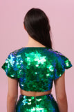 The back of a woman with brown hair, wearing a festival sequin cape made with small and large round holographic Rosa Bloom sequins. The Emerald sequin cape, sitting neatly over her shoulders in this Inti design glistens in the light, creating a mix of shimmering colours of green and blue. The model is also wearing a matching sequin vest top and sparkly leggings.