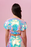 A rear view of a woman long brown hair, swept back to the side, wearing an opal festival sequin cape made with small and large round holographic Rosa Bloom sequins. The Opal sequin cape in this Inti design glistens in the light, creating a mix of shimmering colours of pinks, blues, purples and whites. The model is also wearing a matching sequin vest top and sparkly leggings.