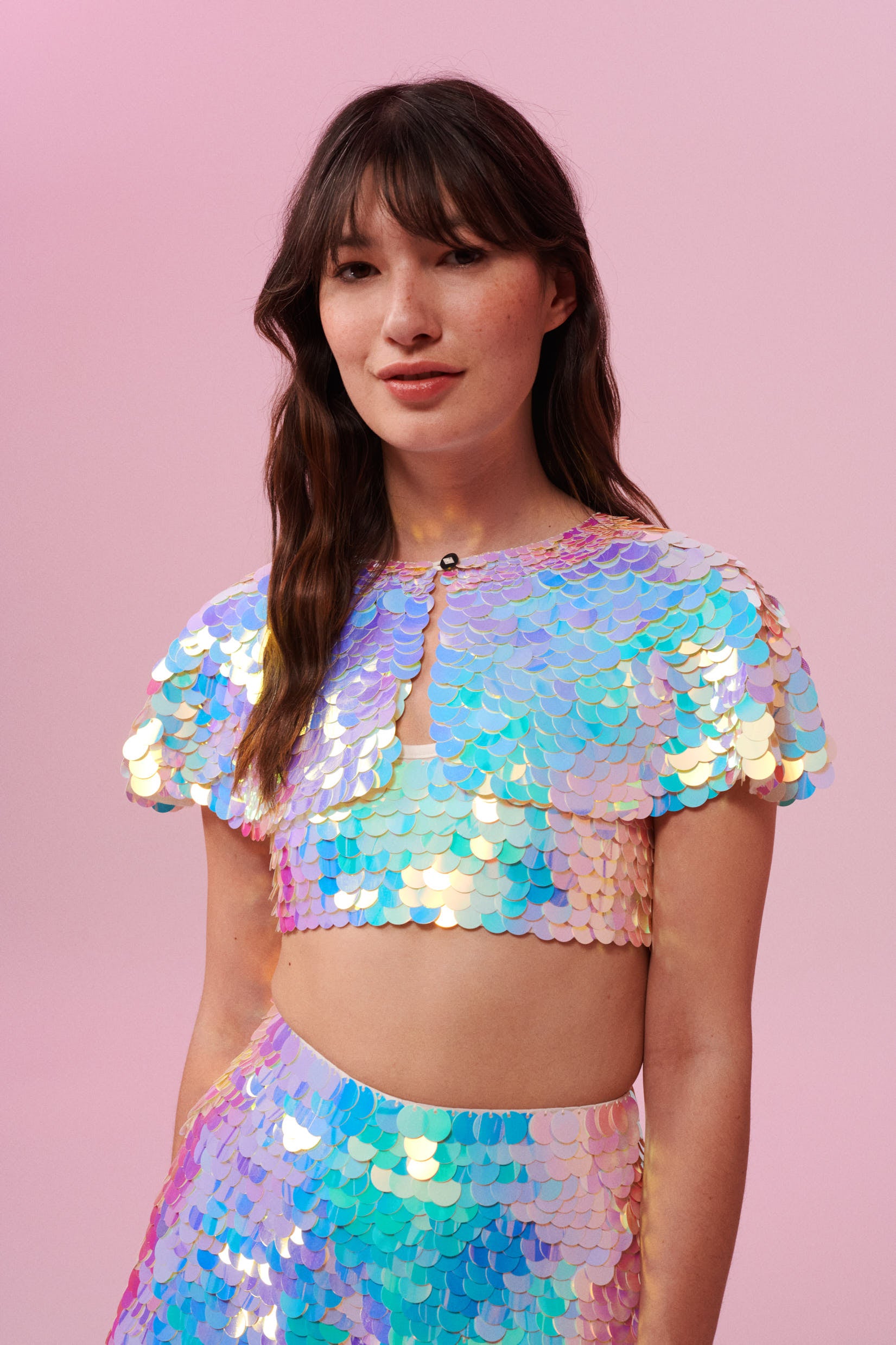 A woman long brown hair with a fringe, swept back to the side, wearing an opal festival sequin cape made with small and large round holographic Rosa Bloom sequins. The Opal sequin cape in this Inti design glistens in the light, creating a mix of shimmering colours of pinks, blues, purples and whites. The model is also wearing a matching sequin vest top and sparkly leggings.