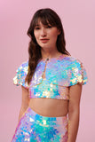 A woman long brown hair with a fringe, swept back to the side, wearing an opal festival sequin cape made with small and large round holographic Rosa Bloom sequins. The Opal sequin cape in this Inti design glistens in the light, creating a mix of shimmering colours of pinks, blues, purples and whites. The model is also wearing a matching sequin vest top and sparkly leggings.