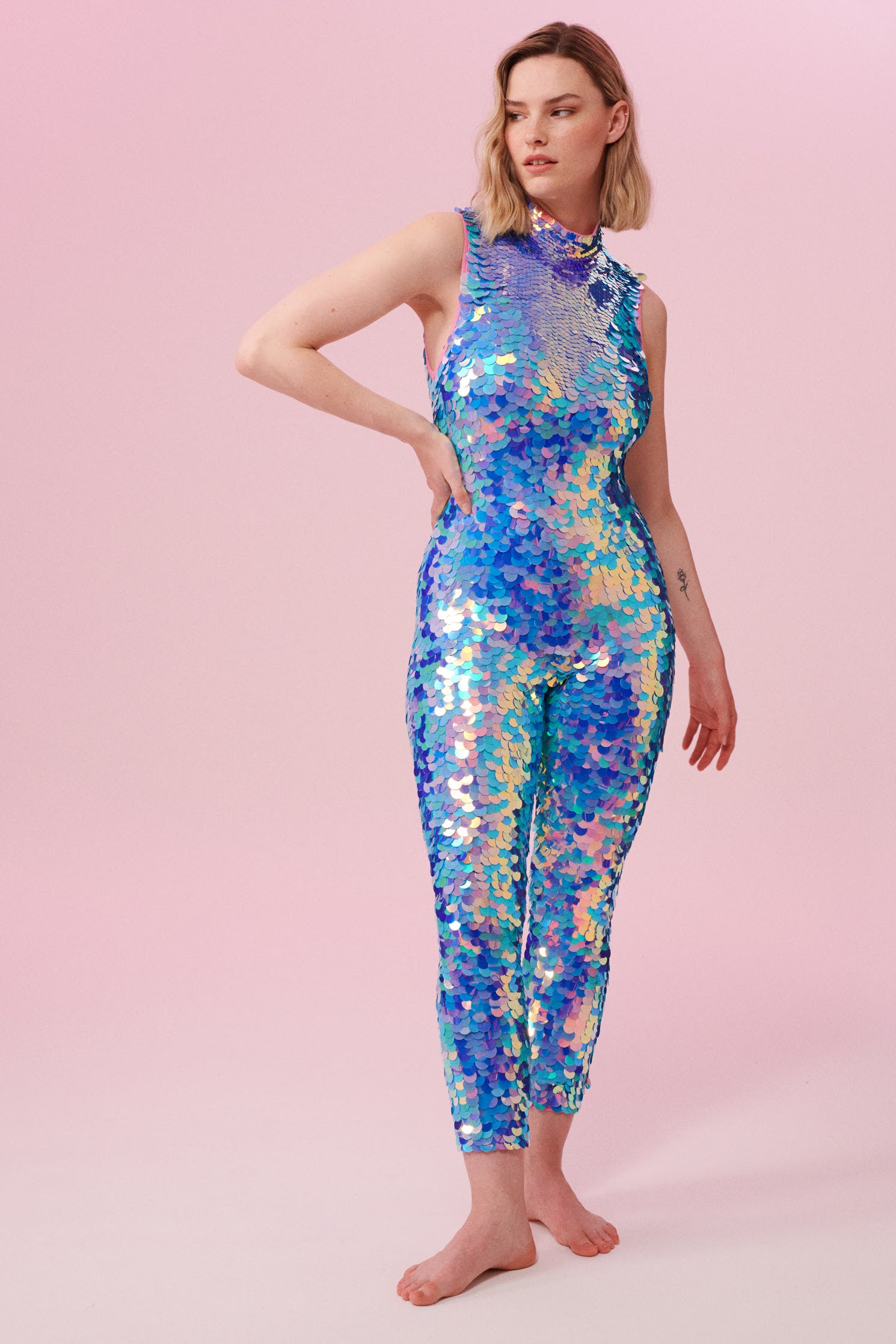  A woman with short blonde hair, wearing an all-in-one sequin stretchy festival jumpsuit made with large round holographic Rosa Bloom sequins. The Iris sequins glisten, creating a mix of shimmering colours of pink, blue and purple.  