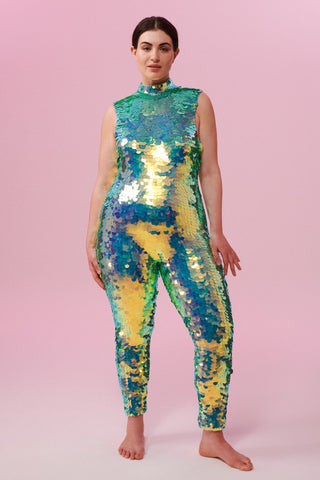 A woman with brown hair, wearing an all-in-one sequin stretchy festival jumpsuit made with large round holographic Rosa Bloom sequins. The Iris jumpsuit sequins glisten all over in this chameleon colour way, creating a mix of shimmering colours of soft mint green and sage that sparkles in the light.